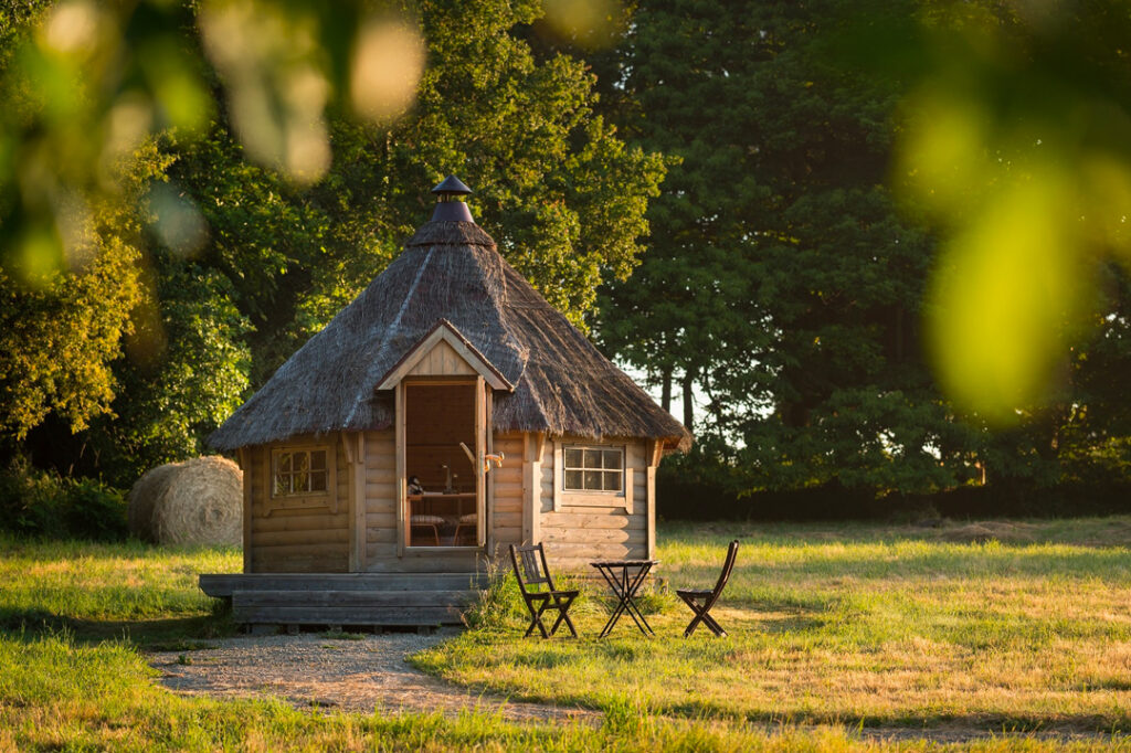 The Lapland cottage at Dihan, an octagonal wooden kota in the meadow at sunset, with a small terrace and two garden chairs